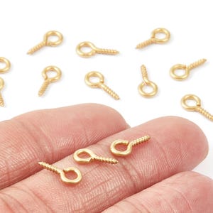 May include: A collection of small, gold-colored screw eye pins. These jewelry-making findings have a screw end and a looped eye for attaching to other components. The pins are scattered on a white surface and held in a hand.