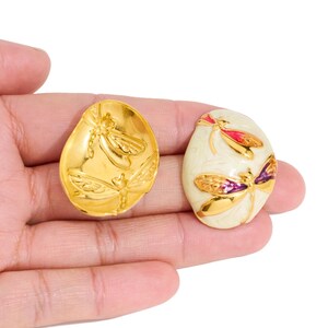 Op de afbeelding: Twee ovale oorbellen. Eén is goudkleurig met een libellenreliëf. De andere is gebroken wit met gouden en paarse libellen details. De oorbellen worden in een hand gehouden tegen een witte achtergrond. Sieraden.
