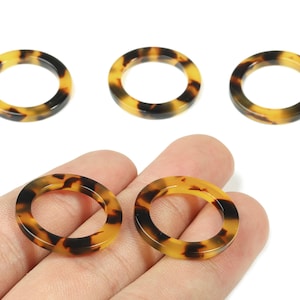 Op de afbeelding: Vijf ronde harsringen met een open centrum en een schildpadpatroon. Twee ringen worden in een hand gehouden, terwijl er drie op een wit oppervlak zijn geplaatst. De ringen hebben bruine, zwarte en geel-oranje tinten.