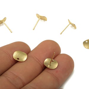 May include: Gold-colored earring posts and backs. The posts have a thin, straight design, and the backs are oval-shaped with a small hole for the post. Several pieces are scattered, with some held in a hand.