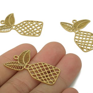 May include: Gold-colored metal charms in a pineapple shape. The charms have a lattice design on the body and a leaf-like top. One charm is held in a hand, while two others are on a white background.