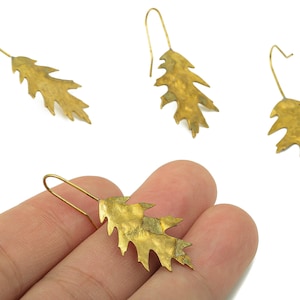 May include: Gold-colored leaf earrings with a hook closure. The earrings are in the shape of oak leaves, with detailed veining and a textured surface. The earrings are approximately 2 inches long.