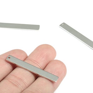 Puede incluir: Abalorios rectangulares de metal plateado con un pequeño agujero en un extremo. Se muestran tres abalorios, uno sostenido en una mano. Estos componentes para la fabricación de joyas son ideales para crear collares, pulseras y pendientes.
