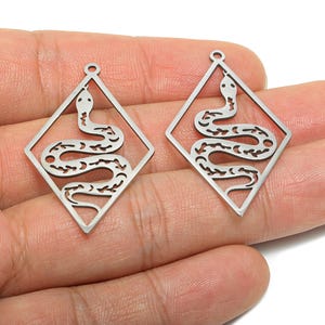 Puede incluir: Dos colgantes plateados con forma de serpiente. Cada colgante presenta un diseño de serpiente dentro de un marco en forma de diamante. La serpiente tiene un patrón detallado. Los colgantes tienen un pequeño lazo en la parte superior para colgar. Estos colgantes son adecuados para la fabricación de joyas.