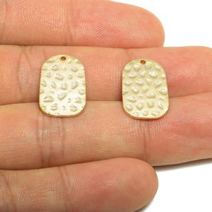 May include: Two gold-colored, textured metal pendant charms. Each charm is rectangular with rounded edges and a small hole at the top. The surface has a pattern of raised, irregular shapes. Jewelry making supplies.