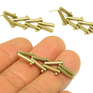 May include: Gold-colored earrings with a geometric design. Each earring features multiple small, cylindrical bars connected with tiny spherical accents. One earring is held in a hand, while two earrings are displayed against a white background.