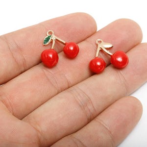 Op de afbeelding: Twee kersvormige bedels met rode, glanzende bollen en goudkleurige stelen en bladeren. De bedels worden in een hand gehouden tegen een witte achtergrond. Deze bedels zijn geschikt voor het maken van sieraden.