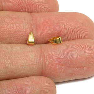 May include: Two small, gold-coloured jewellery findings are displayed. The findings have a rounded, rectangular shape with a small loop at the top. The findings are likely used for jewellery making, such as connecting chains or adding charms.