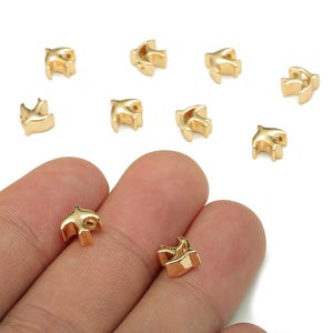 Könnte beinhalten: Kleine, goldfarbene Vogelanhänger. Mehrere Anhänger sind auf einer weißen Oberfläche verstreut, einige werden in der Hand einer Person gehalten. Die Anhänger haben ein dreidimensionales Design und eine glatte, metallische Oberfläche.