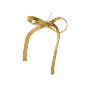 May include: A gold metal bow with a textured surface. The bow is tied with a single knot and has a small, round bead in the center.