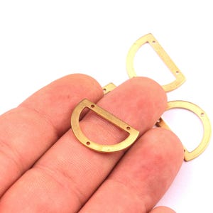 May include: Several small, gold-colored metal D-rings with three holes each are displayed against a white background. The D-rings are held in a person's hand. The rings have a flat top and rounded bottom.