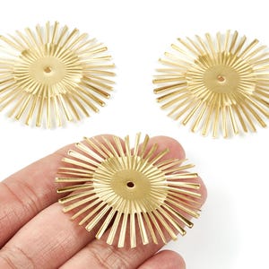 May include: Three gold-colored, sunburst-shaped metal components. Each component has a central circular piece with radiating lines, and a small hole in the center. One is held in a hand.