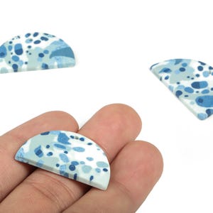 Op de afbeelding: Drie halfcirkelvormige knutselkralen met een terrazzopatroon in blauw- en wittinten. Eén kraal wordt in een hand gehouden, met twee andere in de buurt verspreid. De kralen hebben een platte achterkant en een licht gebogen voorkant.