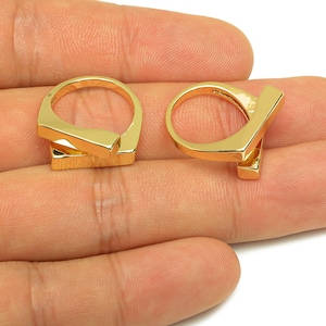 Könnte beinhalten: Zwei goldfarbene Ringe mit geometrischem Design. Jeder Ring hat einen rechteckigen Balken, der das kreisförmige Band schneidet. Die Ringe werden auf einer Hand präsentiert und zeigen ihre polierte, reflektierende Oberfläche.