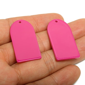 Puede incluir: Dos colgantes de pendientes rosas brillantes en forma de rectángulo arqueado. Cada colgante tiene una parte superior redondeada con un pequeño agujero para la fijación. Los colgantes son lisos y brillantes, y se sostienen en una mano.