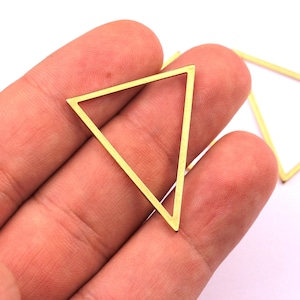 Peut inclure: Une main tient un triangle doré à cadre ouvert. Une autre forme dorée à cadre ouvert, un carré, est visible en arrière-plan. Ces formes géométriques sont probablement des composants pour la fabrication de bijoux ou des projets d'artisanat.
