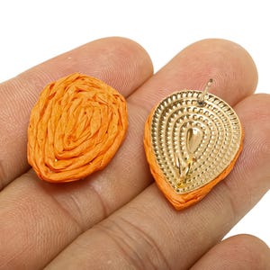 May include: Close-up of two orange raffia earrings. One is a round, spiral design, and the other is a teardrop shape with a gold-coloured metal backing. The earrings are held in a person's hand.