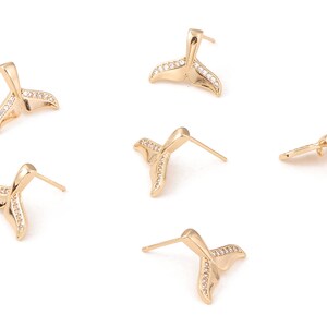 Puede incluir: Pendientes de botón dorados con forma de cola de ballena. Cada cola está adornada con pequeños cristales transparentes a lo largo de los bordes. Los pendientes tienen un poste y un cierre para un uso seguro.