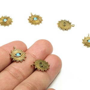 May include: Gold-colored sunburst charms with a central turquoise-colored eye design. The charms have a small loop at the top for attaching to jewelry. The charms are small, with a detailed sunburst pattern around the eye.
