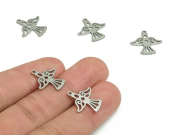 Puede incluir: Varios pequeños dijes plateados con forma de ángel, con alas caladas y faldas fluidas. Los dijes están dispersos sobre un fondo blanco, algunos se sostienen en una mano. Los dijes son de metal y tienen un pequeño agujero en la parte superior.