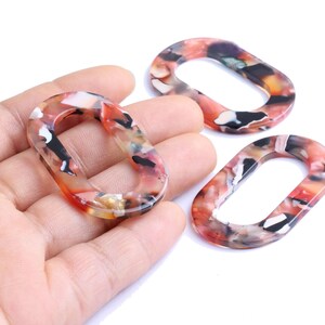 Peut inclure: Trois maillons ovales en acrylique avec un motif marbré rouge, noir, blanc et orange. Un maillon est tenu dans une main, les deux autres sont sur une surface blanche. Ces maillons sont adaptés à la création de bijoux.