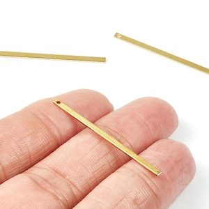 May include: Three gold-coloured, rectangular bar charms with a small hole at one end. One charm is held in a hand, while the other two are positioned on a white background. These charms are ideal for jewellery making.