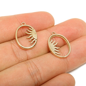 May include: Two gold-colored sun-shaped pendants. Each pendant features a sun design within an oval frame. The sun has a face and rays. The pendants are small and have a loop at the top for hanging.