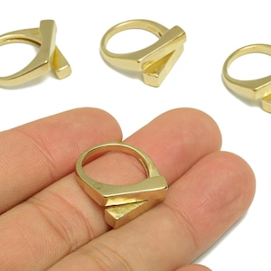 May include: Gold-colored rings with a geometric design. The rings have a rectangular bar that intersects with the ring band. One ring is on a person's hand, and three others are on a white background.