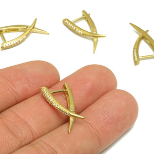Pode incluir: Brincos dourados com um design cruzado único. Um dos brincos é segurado na mão, mostrando os detalhes intrincados. Os brincos apresentam uma barra curva com pequenos detalhes brilhantes e uma ponta.