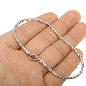 Puede incluir: Una pulsera plateada con un diseño de cadena trenzada. La pulsera tiene un cierre de gancho y se muestra en una mano.