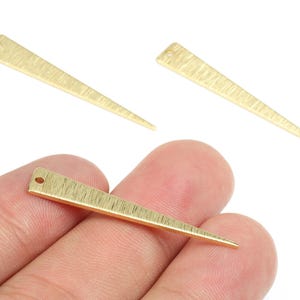 May include: Three gold-colored, textured metal triangle charms. One charm is held in a hand, while two others are positioned above. Each charm has a small hole at the top.