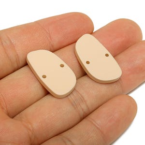 May include: Two beige, half-oval shaped beads with two small holes each. The beads are held in a person's hand against a white background. These beads are likely used for jewellery making or other craft projects.
