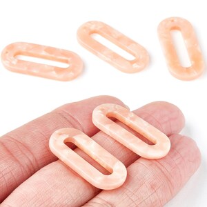 May include: Five peach-coloured oval acrylic beads with a marbled effect. Each bead has a central hole. These beads are suitable for jewellery making and crafting projects. The beads are shown in a hand.