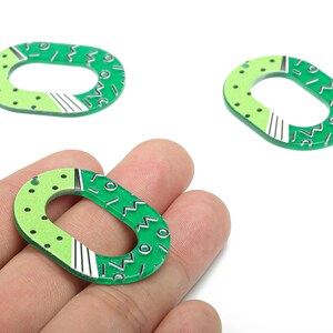 May include: Oval green acrylic charms for jewellery making. The charms have a light green section with black dots and a dark green section with white and black geometric designs. The charms are held in a hand.