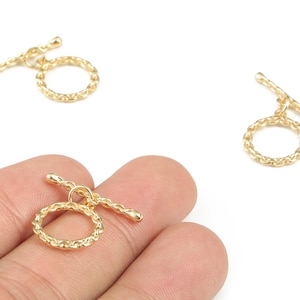 May include: Gold-coloured toggle clasps, suitable for jewellery making. The clasps feature a textured ring and a straight bar. One clasp is held in a hand, while two others are scattered on a white background. These clasps are perfect for necklaces and bracelets.