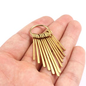 May include: A gold-colored, fan-shaped pendant with multiple thin, rectangular bars radiating from a central ring. The pendant is held in a person's hand against a white background. The bars are of varying lengths.