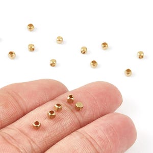 May include: Small, gold-colored metal beads scattered on a white surface, with a few held in a person's fingers. The beads are hexagonal in shape and have a hole running through the center. These beads are suitable for jewelry making and crafts.