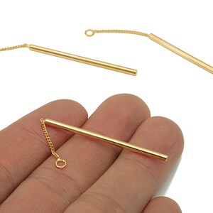 May include: Gold-colored jewelry findings, including three straight bar pendants with attached delicate chains and small loops. One pendant is held in a hand. The findings are likely for earrings or necklaces.