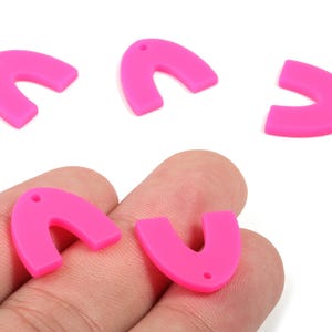Pode incluir: Pingentes de acrílico rosa brilhante em forma de U, com um pequeno orifício para enfiar. Vários pingentes estão espalhados em um fundo branco, com dois sendo segurados em uma mão. Ideais para fazer joias ou projetos de artesanato.