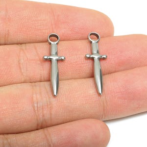 Peut inclure: Deux petits charmes en forme de dague de couleur argentée. Chaque breloque a une boucle en haut, une garde transversale et une lame pointue. Les breloques sont présentées sur un fond neutre.
