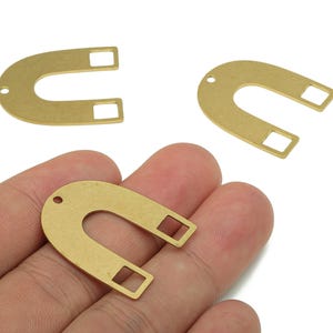 May include: Three brass-colored horseshoe-shaped charms with rectangular cutouts at the ends. One charm is held in a hand, while the other two are scattered in the background. Each charm has a small hole at the top.