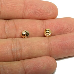 May include: Two small, gold-colored beads with a ribbed design. The beads are spherical and have a shiny finish. The beads are shown on a person's hand.