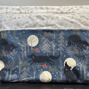 May include: A blue zippered pouch with a white and gray patterned top. The bottom of the pouch features a repeating pattern of black wolves howling at the moon in a dark blue forest.