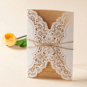May include: A white wedding invitation featuring a delicate lace design. The invitation is tied with a brown twine bow and has a kraft paper insert. The text on the insert reads "Together with our families".