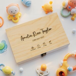 May include: A light wooden keepsake box engraved with the name "Amelia Rose Taylor" and birth details. The box is surrounded by colorful baby toys, including rattles and pacifiers.