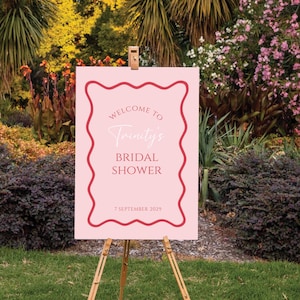 May include: A rectangular light pink sign on a wooden easel featuring a red wavy border. The text reads: Welcome to Trinity's Bridal Shower, 7 September 2029. The sign is displayed outdoors in a garden setting with various green plants and flowers.