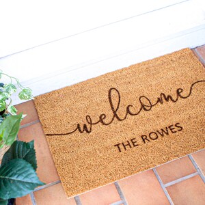 May include: A brown coir doormat with the text "welcome" in a cursive font and "THE ROWES" in a block font.
