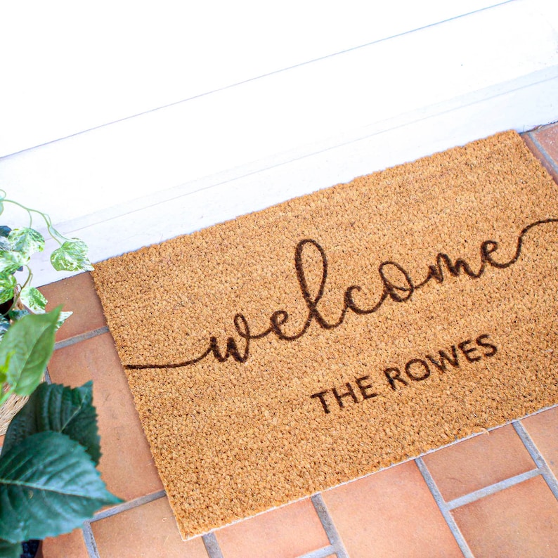 May include: A brown coir doormat with the text "welcome" in a cursive font and "THE ROWES" in a block font.
