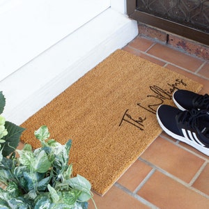 May include: A brown coir doormat with the text "The Williams" in black cursive lettering. The doormat is placed on a brick patio.
