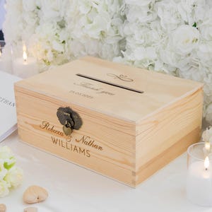 May include: A light-colored wooden box with a slot on top, likely for cards or gifts. The box features engraved text that reads "Thank you" and a date. It has a decorative clasp and the names "Rebecca & Nathan WILLIAMS" are also engraved on the front.